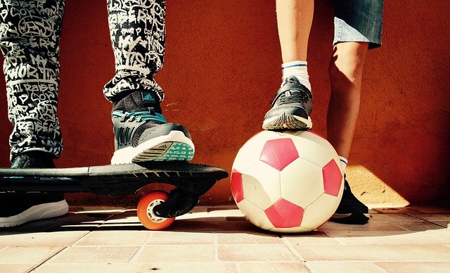 foto van de voeten van jongeren. Eén heeft het been op een skateboard, de ander op een voetbal.